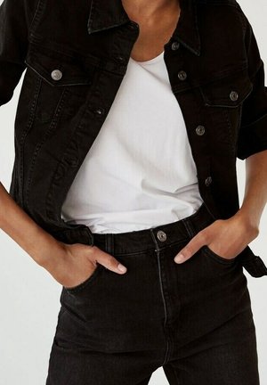 Personne portant une veste en jean noire et un jean avec un t-shirt blanc, les mains dans les poches avant, debout devant un fond uni.