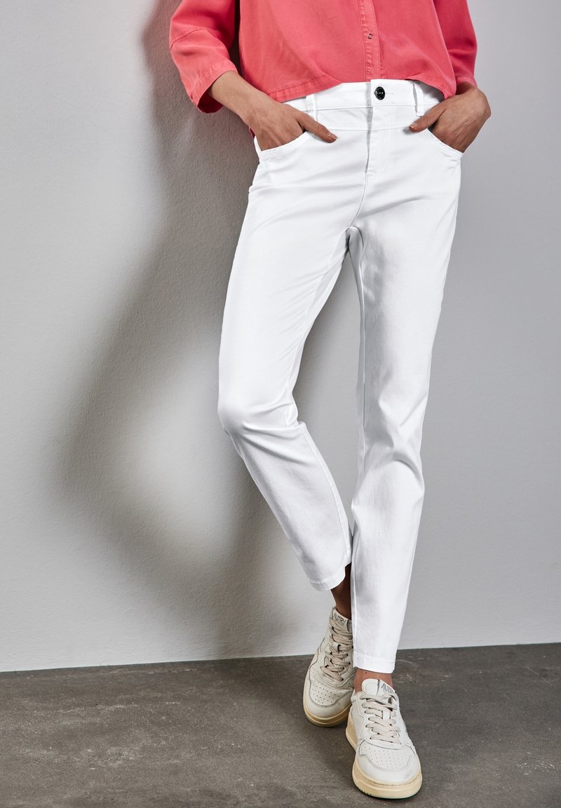 Personne portant un pantalon blanc, des baskets beiges et une chemise corail à manches longues, les mains dans les poches, debout contre un mur gris.