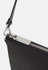 Pochette noire texturée avec une fermeture éclair argentée, dotée d'une sangle élégante et d'un détail de logo sur le matériel. Forme rectangulaire compacte.