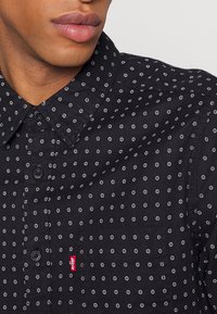 Gros plan d'une personne portant une chemise sombre à boutons avec un petit motif circulaire blanc et une étiquette rouge Levi's sur la poche poitrine.