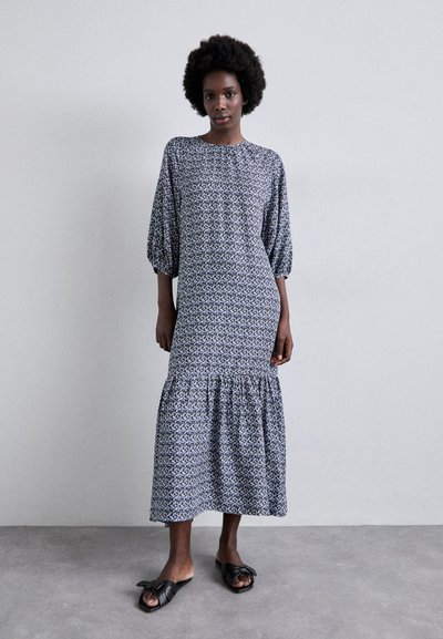 Robe longue à manches bouffantes avec un ourlet en cascades. Tissu à motifs bleu et blanc, coupe ample, portée avec des sandales à glisser noires. Texture lisse.