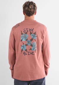 Chemise en coton rose à manches longues avec un motif floral bleu et marron, portant le texte "STAY ALIVE" au dos, col rond et coupe décontractée.