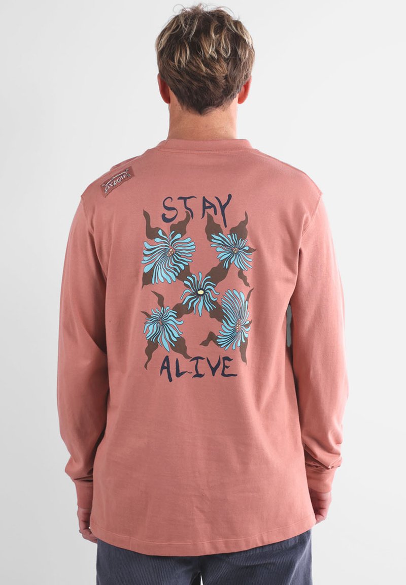 Chemise en coton rose à manches longues avec un motif floral bleu et marron, portant le texte "STAY ALIVE" au dos, col rond et coupe décontractée.