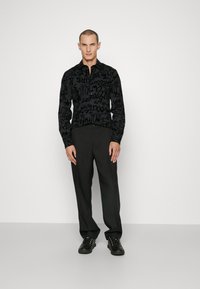 Camicia nera a maniche lunghe con un motivo grafico testurizzato, abbinata a pantaloni scuri e scarpe nere. L'outfit presenta un design slim fit.