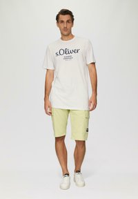 Witte katoenen T-shirt met marineblauwe opdruk "s.Oliver," in combinatie met lichtgele cargoshorts met zijzakken en een logopatch. Witte sneakers.