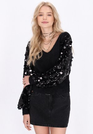 Pull noir avec des manches pailletées, présentant un décolleté en V et une texture douce, associé à une jupe en denim noire.