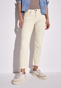 Jeans dritti color crema con orlo sfrangiato, abbinati a sneaker bianche con dettagli beige e rosa. Giacca di denim sovrapposta.