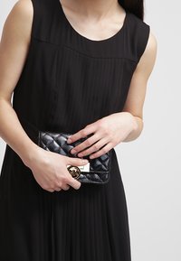 Borsa a pochette nera trapuntata con accentuazione in oro del logo MK, tenuta da una persona che indossa un abito nero senza maniche. Texture liscia, forma compatta.