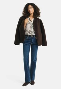 Brauner oversized Mantel, lichtblaues Hemd mit floralem Muster und blaue Jeans mit geradem Bein. Braune Stiefeletten runden das Outfit ab.