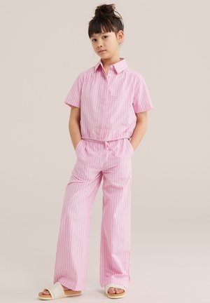 Noor tüdruk seisab käed taskutes, seljas roosa-valgete triipudega lühikeste varrukatega särk ja sobivad laia säärega püksid, jalas beežid sandaalid.