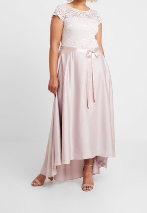 Frau trägt ein hellrosa Spitzenkleid mit einem Satin-Schleifengürtel und hält den fließenden asymmetrischen Rock, kombiniert mit beigen Sandalen mit Absatz.