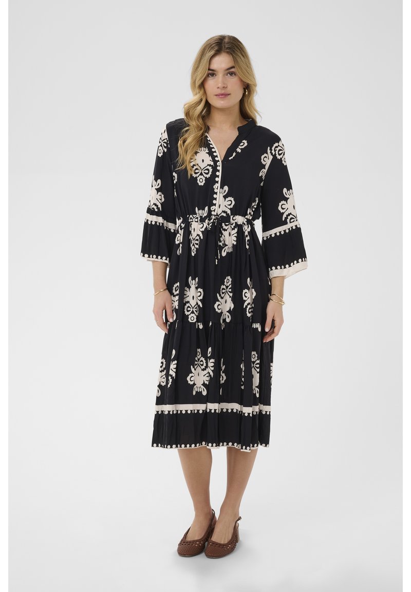 Woman wearing a black midi dress with cream floral patterns, 3/4 sleeves, a drawstring waist, and brown woven flat shoes.