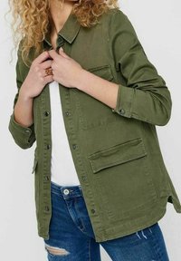 Giacca verde oliva in tessuto canvas, con chiusura a bottoni, maniche lunghe, due tasche sul petto e un fit rilassato.