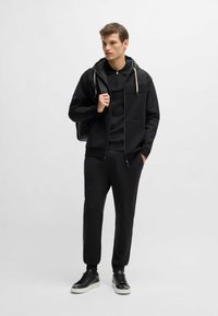 Ensemble de sport noir comprenant un hoodie zippé avec cordons de serrage, un pull assorti et un pantalon fuselé. Des baskets noires complètent le look.