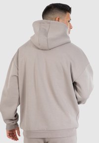Mann mit kurzen dunklen Haaren, der einen übergroßen, hellgrauen Kapuzenpullover trägt und mit dem Rücken zur Kamera vor einem schlichten Hintergrund steht.