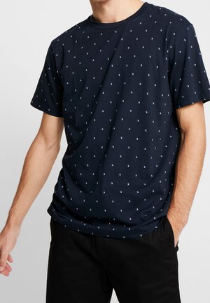 Man wearing navy blue short-sleeve t-shirt with small white and blue geometric pattern, paired with black pants, standing against plain background.