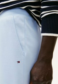 Un pantalon de survêtement bleu clair présente une poche latérale, une taille côtelée et un petit logo brodé en rouge, blanc et bleu près de la poche.
