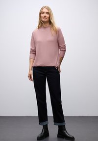 Rosa Pullover mit hohem Ausschnitt und seitlichem Kordelzug, kombiniert mit dunklen Jeans mit hochgekrempelten Säumen und schwarzen Stiefeletten.