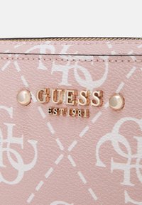 Rosa läderyta med vit monogramdesign och gyllene GUESS-logotyp med "EST 1981" mellan två gyllene nitar.