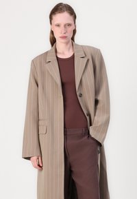 Beige pinstripe coat with a notched collar, single button closure, and two side pockets, worn over a brown top and brown trousers.