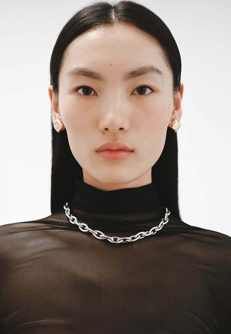 Collar de cadena de plata con eslabones circulares, usado sobre un cuello de tortuga negro y transparente. La modelo lleva pendientes geométricos de oro y tiene el cabello negro y liso.