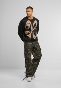 Schwarzer Pullover mit einer großen beigen Grafik in der Mitte, kombiniert mit dunkel-tonierten Cargo-Hosen im Camouflage-Muster und weißen Sneakers.