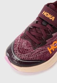 Beh spustových topánok HOKA v odtieňoch tmavočervenej a ružovej. Sieťovaná vrchná časť s textúrovaným vzorom a oranžovým logom. Hladká, lesklá špička a odpružená podrážka.