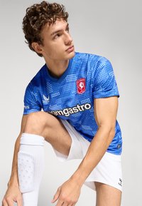 Modrý fotbalový dres s tečkovaným vzorem, krátké rukávy a výstřih do V. Obsahuje logo na hrudi a bílé šortky s bílými ponožkami do kolen.