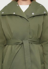 Kvinna iklädd en olivgrön belagd kappa med stor krage och silverknappar, över en vit topp, sedd från halsen till höften.