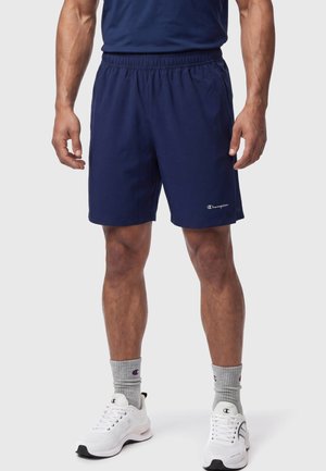 Mann trägt dunkelblaue Champion-Shorts, graue Champion-Socken und weiße Champion-Sneaker und steht vor einem schlichten Hintergrund.