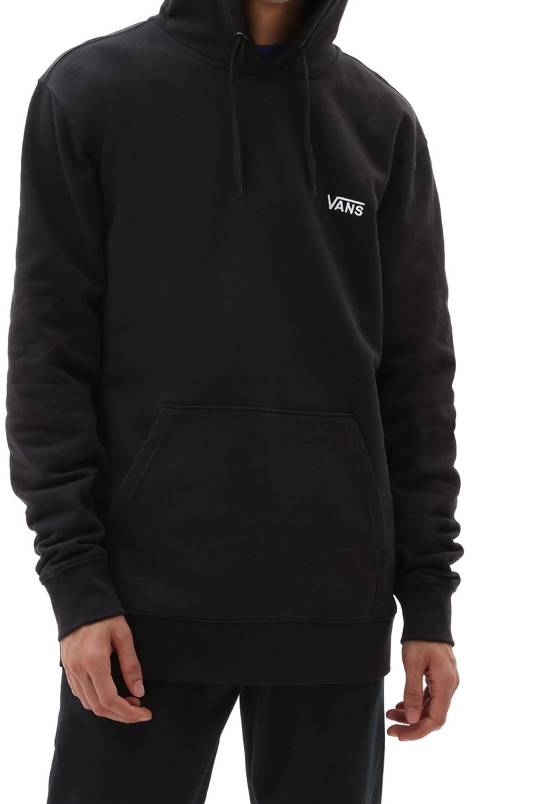 Personne portant un sweat à capuche noir Vans avec une poche avant et un logo sur la poitrine, debout devant un fond blanc.