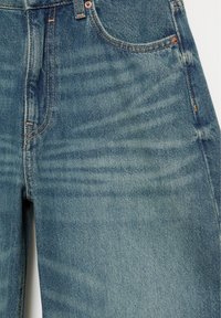 Gros plan sur un jean en denim bleu montrant les poches avant, les passants de ceinture et les détails de couture orange sur un tissu texturé.