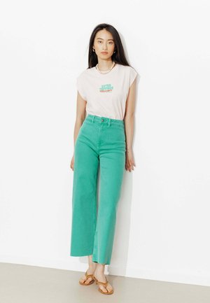 Jeune femme aux longs cheveux foncés portant un haut blanc sans manches avec un texte, un jean taille haute couleur sarcelle et des sandales marron, debout contre un mur blanc.