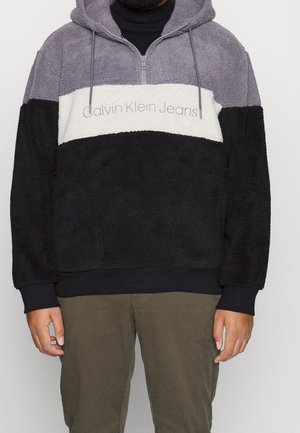 Mand iført en color-blocked fleece-hoodie med grå, hvide og sorte sektioner og "Calvin Klein Jeans"-logo på tværs af brystet.