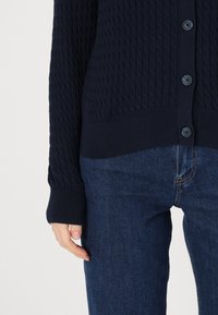 Μπλέ πλεχτό ζακέτα με ανάγλυφο σχέδιο καλωδίων, ολοκληρωμένη με κουμπιά και ριμπ μανσέτες. Φοριέται πάνω από σκούρα μπλε τζιν.