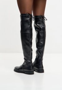 Botas de cuero negras hasta el muslo con una textura suave, con lazos traseros y un zipper lateral para poner y quitar fácilmente. Suelas planas y robustas.
