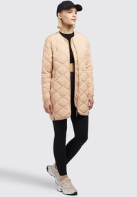 Chaqueta larga acolchada en un tono durazno claro con patrones de diamantes redondeados, cierre frontal, bolsillos laterales, combinada con leggings negros y zapatillas beige.