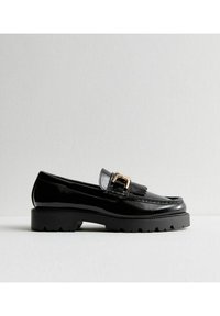 Mocasines de piel sintética negra con suela de goma gruesa, que presentan una hebilla decorativa dorada y un detalle con flecos en la parte frontal.