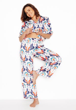 Femme souriante levant un haut de pyjama à motifs, portant un pantalon assorti avec des dessins colorés d'oiseaux et de fleurs, debout pieds nus sur une jambe.
