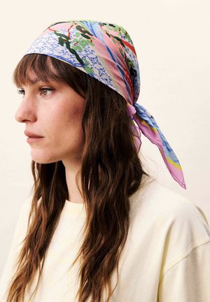 Femme aux longs cheveux bruns portant un foulard coloré à motifs floraux et de script noué à l'arrière, haut beige, tournée vers la gauche.