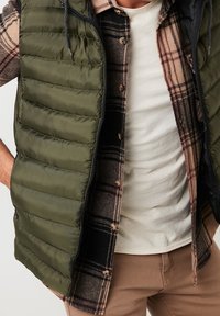 Olivově zelená důlková vesta přes kostkovanou košili s hnědými, krémovými a černými tóny; hladká textura s viditelným šitím a zipem.