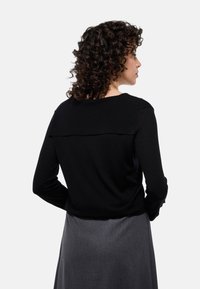 Haut noir à manches longues avec une texture côtelée et un dos en couches. Porté avec une jupe grise, montrant un fini en tissu lisse.