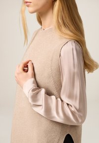 Beiger gestrickter ärmelloser Pullover mit gerippten Rändern, getragen über einer hellrosa Bluse. Verfügt über Seitenschlitze und eine glatte Textur.