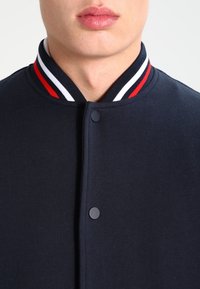 Marine bomberjack met een opstaande kraag, voorzien van een rood, wit en zwart gestreept kraag en een drukknoopsluiting aan de voorkant.