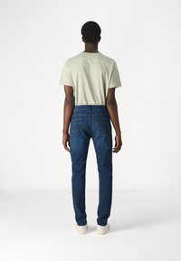 Jeans in denim blu scuro con taglio slim, dotati di tasche posteriori e dettagli di cucitura sottili. Abbinati a una camicia a maniche corte di un verde chiaro.