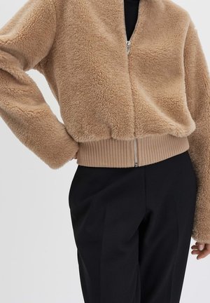 Chaqueta beige borrosa con cremallera y dobladillo acanalado, usada con pantalones negros, mostrando una mano en la cadera y un ajuste relajado.