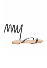 Sandali neri piatti con sottili cinturini e un design unico a spirale alla caviglia. La soletta è beige e piatta, offrendo un aspetto minimalista.