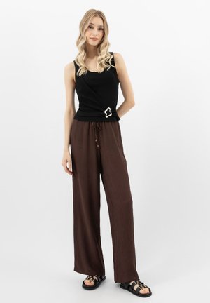 Blonde vrouw met een mouwloze zwarte top met een gespdetail, losse bruine broek en zwarte sandalen, staand tegen een effen witte achtergrond.