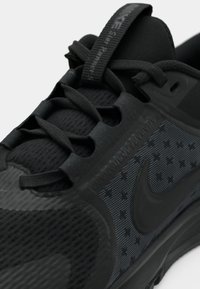 Svart idrettssko med teksturert overflate, mesh-detaljer og et fremtredende merke. Har flate lisser og en polstret såle.