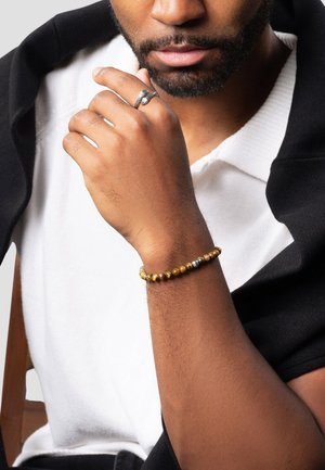 Perlekæde med runde perler i guld, sølv og brun, båret på et håndled. Glat tekstur, på en lys baggrund.